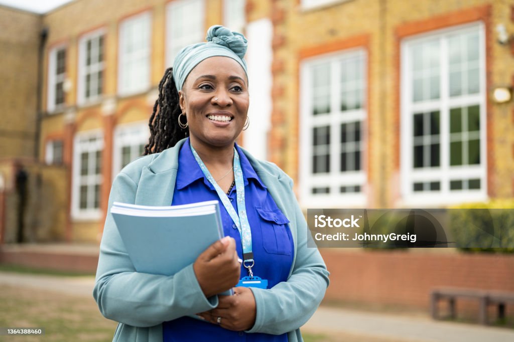 Cheerful Black teacher standing outside education building Candid portrait of smiling early 50s woman in businesswear and headscarf holding stack of composition booklets and looking away from camera. Teacher Stock Photo