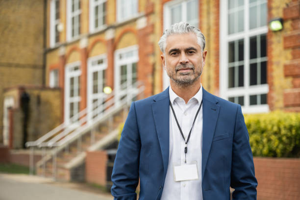 Multiracial male principal standing outdoors on campus Waist-up front view of early 50s grey-haired man wearing button down shirt, blue suit jacket, ID lanyard, and looking at camera with confident smile. high school teacher stock pictures, royalty-free photos & images