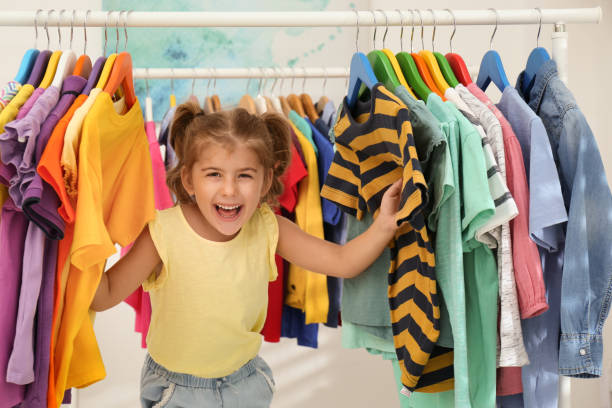 забавная маленькая девочка, выбирающая одежду на вешалке в помещении - child clothing arrangement hanger стоковые фото и изображения