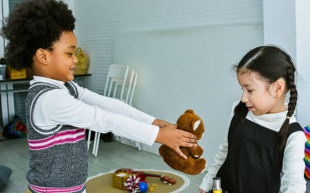 愛らしいアフリカの黒い小さな男の子は、自宅で快適なリビングルームに立って、共有し、かわいい女の子に人形や贈り物を与え、幸せで微笑みます。愛、多様性、子供、教育コンセプト。 - 分かち合い ストックフォトと画像