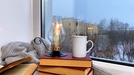 Coziness winter atmosphere. Coffee cup with smoke, candle burning and books on windowsill, winter morning or evening, snowing outside, wind moving pages. Selective focus