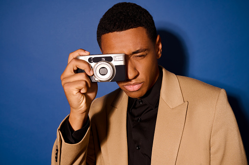 Photographing. Confident dark-skinned guy with closed eye in light suit holding camera with one hand near his face standing on blue background in studio