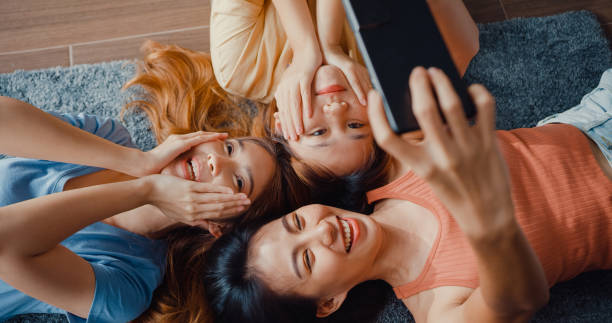 Above view close up group of Asia ladies with happiness enjoy moment hold smartphone smiling take memories picture lying on carpet in floor living room at home. Lifestyle activity quarantine concept. Above view close up group of Asia ladies with happiness enjoy moment hold smartphone smiling take memories picture lying on carpet in floor living room at home. woman lying on the floor isolated stock pictures, royalty-free photos & images