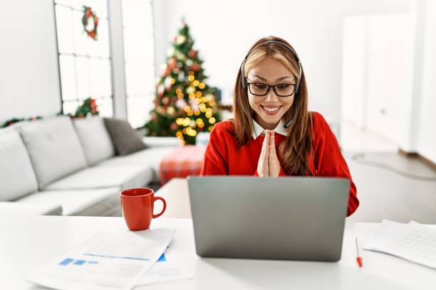 istock Young caucasian girl sitting on the table working using laptop by christmas tree praying with hands together asking for forgiveness smiling confident. 1364404991