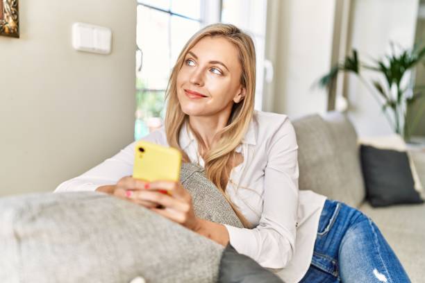 istock Young blonde woman using smartphone sitting on the sofa at home. 1364409024