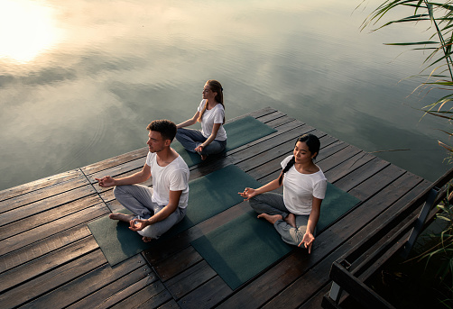 Group of people doing yoga exercises by the lake at sunset.