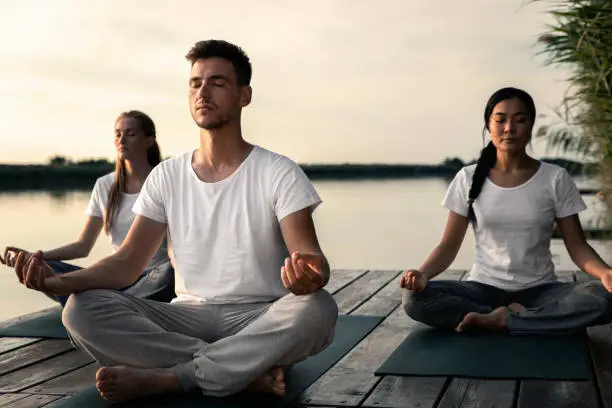 Photo of Group of people doing yoga exercises by the lake at sunset.