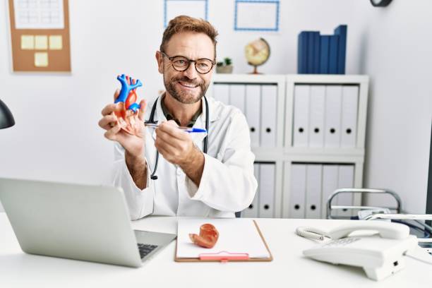 Middle age hispanic man wearing doctor uniform holding heart at clinic stock photo