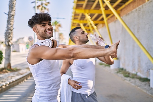 Two hispanic men couple smiling confident stretching and drinking water at street