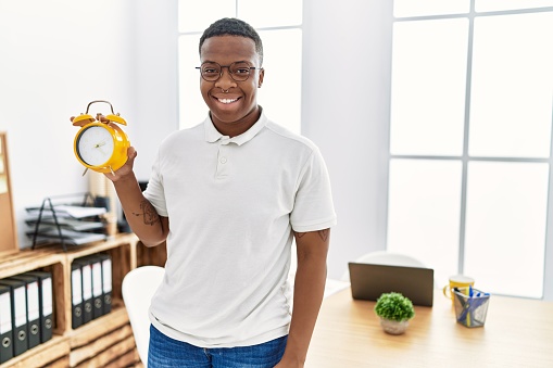 Young african man holding alarm clock at the office looking positive and happy standing and smiling with a confident smile showing teeth