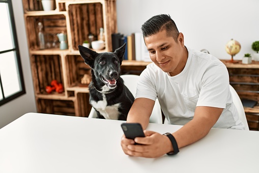 Young hispanic man smiling happy using smartphone sitting on the table with dog at home.