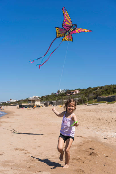 kleines mädchen, das während einer reise mit der familie mit fliegendem drachen am großen einsamen strand läuft - kleines mädchen, das an einem sonnigen frühlingstag mit drachenschmetterling an der küste spielt - color image travel locations sports and fitness nature stock-fotos und bilder