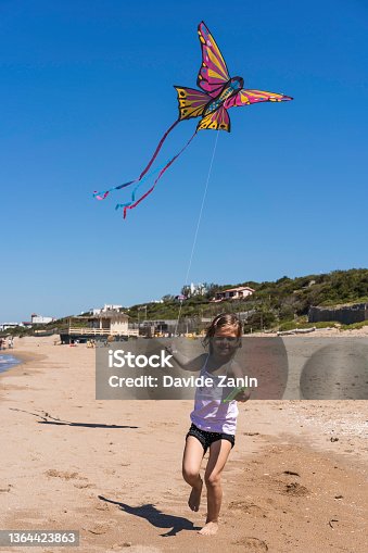 istock Little girl running with flying kite on the big deserted beach during a travel with family - Little girl playing with kite butterfly on sea shore in a spring sunny day 1364423863