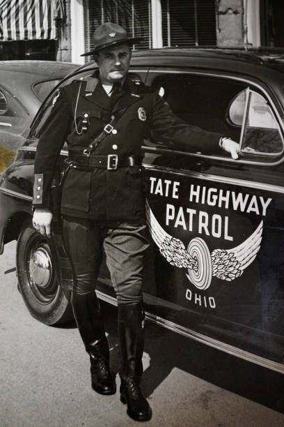 Lima, Ohio, USA - July 20, 1946: Ohio state highway patrolman Joseph Ballard standing by 1946 Ford  patrol car, 1 person, black and white image, close view, standing with hand on patrol car. Image taken by James Ballard of Springfield, Ohio,   The Ohio State Highway Patrol was founded in 1933 . Archival photo owned by Jerry Ballard.