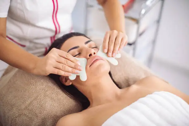 Beautiful mid adult woman receiving face massage with jade stones. Gua sha is traditional Chinese massage technique for relaxation and beauty and body care