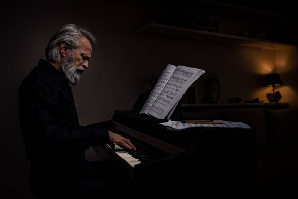 arrangiato dai capelli grigi, barbuto, affascinante compositore, pianista, artista di mezza età in una camicia nera a casa in una stanza buia con lampada ambientale e luce, concentrato, impilando e suonando il pianoforte elettrico in mezzo, bevendo una ta - piano men pianist musician foto e immagini stock