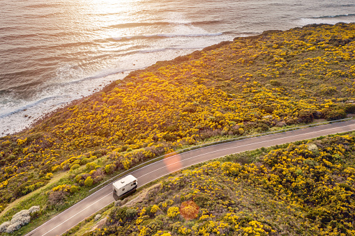 Driving a motor home on a road by the sea at sunset, with warm atmosphere and lens flare, seen from the air. Beautiful coastal scenery with sea waves breaking on a beach. Camping life, alternative lifestyle, freedom.