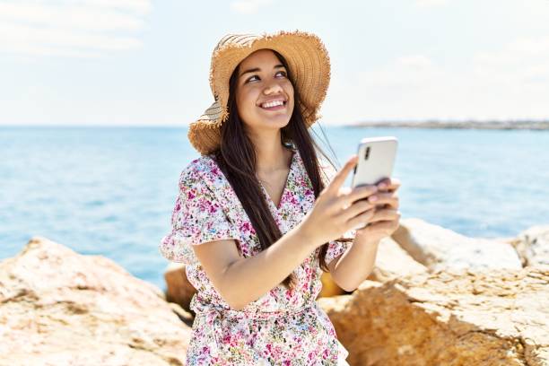 istock Young latin girl wearing summer hat using smartphone at the beach. 1364444455