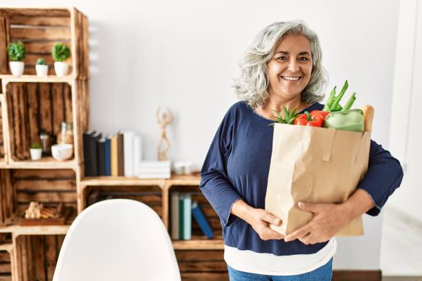 Middle age grey-haired woman holding paper bag with groceries standing at home. stock photo
