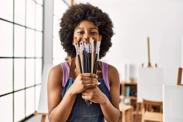 Young african american woman covering mouth with paintbrushes at art studio stock photo