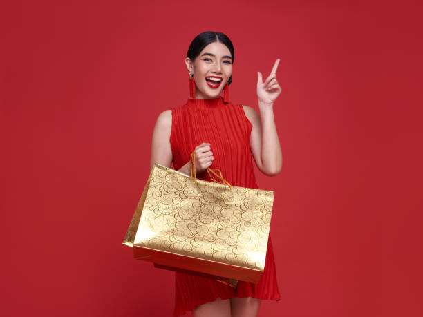 Smiling beautiful Asian woman holding shopping bags and pointing to copy space isolated on red background for Chinese new year sale concept. stock photo