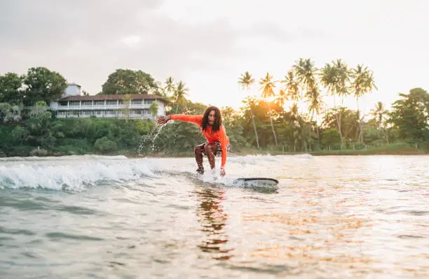 Photo of Black long-haired teen boy riding a long surfboard. He caught a  wave in an Indian ocean bay with magic sunset background. Extreme water sports and exotic countries concept.