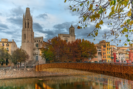 Autumn evening landscape of Girona with Sant Feliu Pedestrian Bridge over Onyar River on the backdrop of Church of St. Felix towers, Spain. Bright cityscape with festive lights reflected in the water