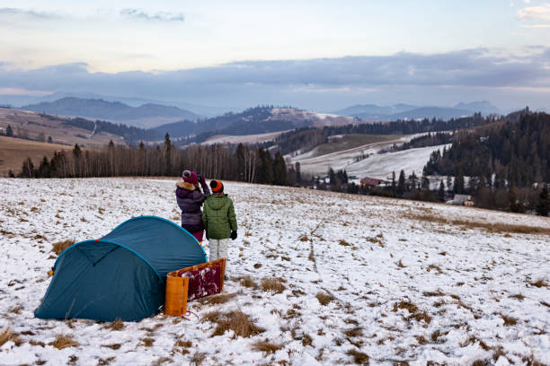 Brother and sister camping on winter hill. They are sitting in tent, drinking hot tea and enjoying looking at winter view.Shot with Canon R5.