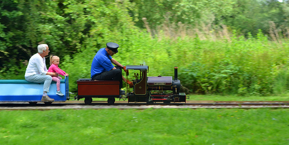 St Neots, Cambridgeshire, England - June 09, 2019: Miniature Steam train giving adults and children rides in the park with driver little girl and grandma.