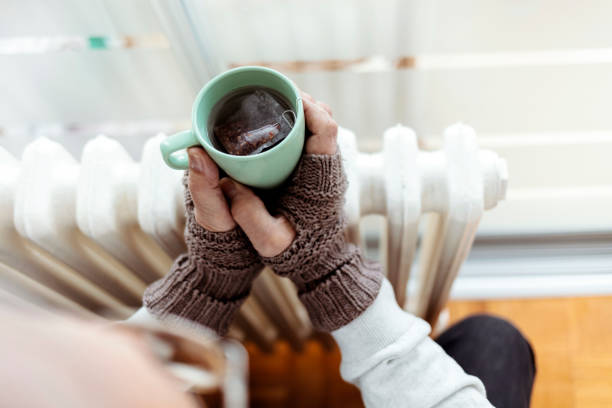 Warming up with hot tea always gives great results. A cold grandfather wearing gloves, warming himself by the radiator and drinking hot tea at home during the day. The hands of an older man holding a hot drink and sitting next to a radiator. Concept of heating season. fuel crisis stock pictures, royalty-free photos & images