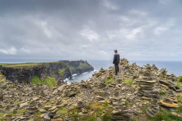Man standing between stone stackings and admiring view on in iconic Cliffs of Moher, Ireland Man standing between stone stackings and admiring view on in iconic Cliffs of Moher, popular tourist attraction, Wild Atlantic Way, Co. Clare, Ireland doolin photos stock pictures, royalty-free photos & images