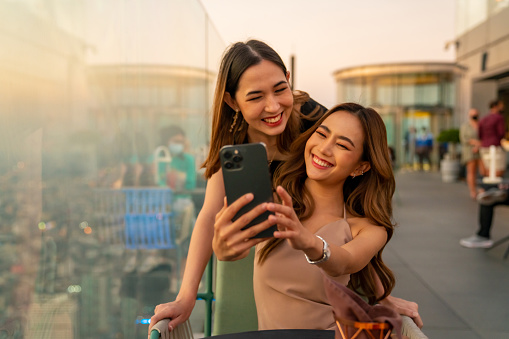 Asian woman friends meeting and using smartphone selfie together at skyscraper rooftop restaurant in metropolis at summer sunset. Female friend enjoy outdoor lifestyle activity in the city at night