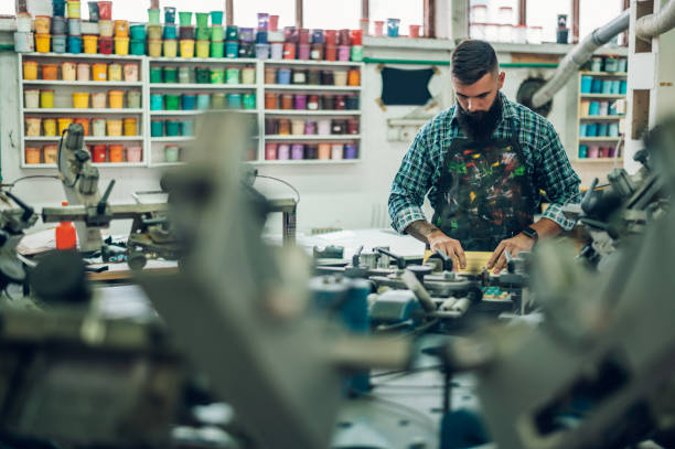 männlicher arbeiter, der tinte auf den rahmen drückt, während er die druckmaschine in einer werkstatt benutzt - silk screen fotos stock-fotos und bilder