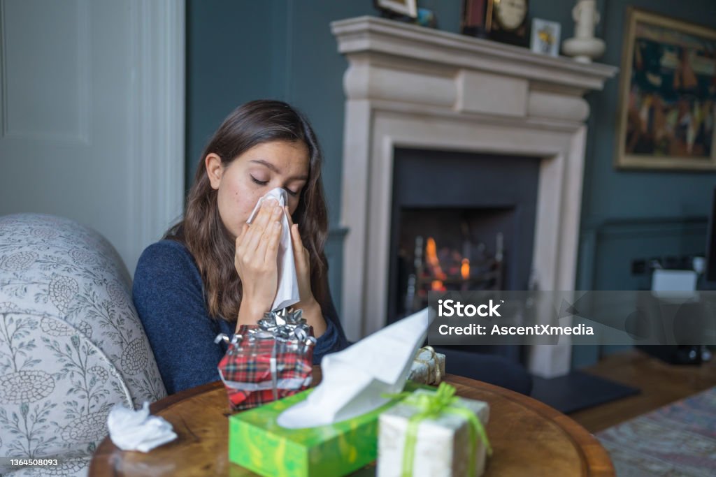 Young woman relaxes in living room, at home She sits beside table of Christmas presents, blowing her nose Christmas Stock Photo