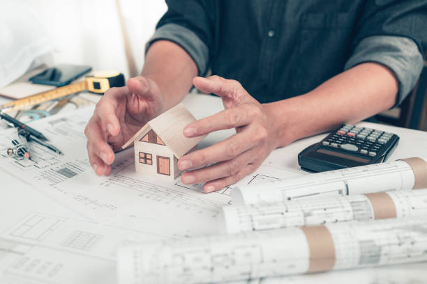 istock Architect engineer hands hold and protect mini house model on blueprint. Protect House, cost planning design and construction concept. 1364534639