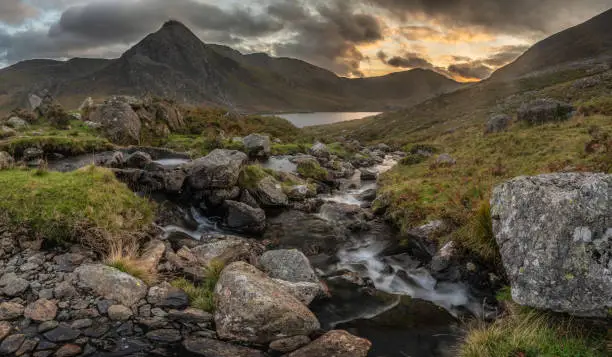 Epic dramatic Autumn sunset landscape image of Llyn Ogwen and Tryfan in Snowdonia National Park with stream and rocks in foreground