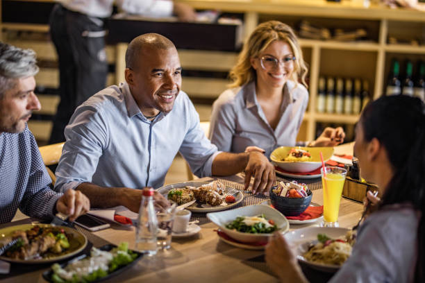 Happy African American entrepreneur talking to his colleagues during lunch break in restaurant. Happy black businessman communicating with his colleagues during lunch in a restaurant. business dinner stock pictures, royalty-free photos & images