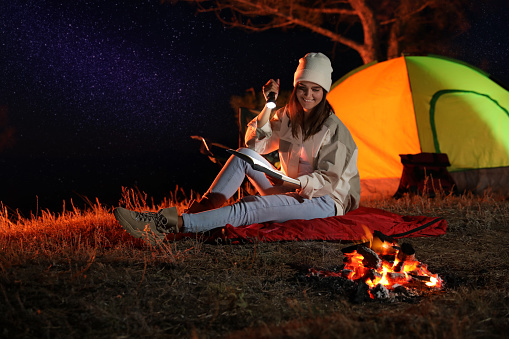 Young woman with flashlight reading book near bonfire at night. Camping season