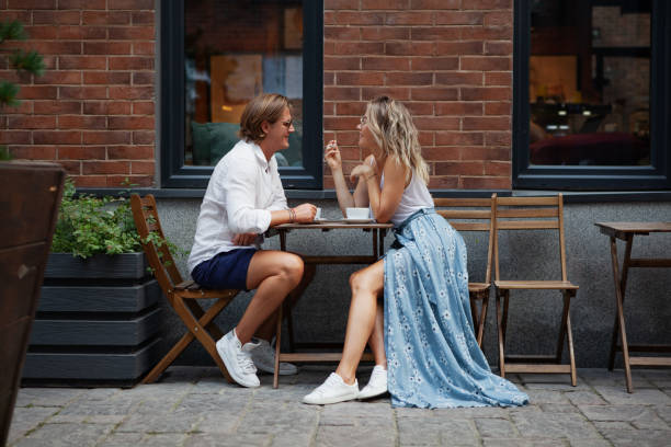 joven pareja de moda hablando y sonriendo en la cafetería en la calle de la ciudad. elegante retrato de hombre y mujer de cuerpo entero en el restaurante. cita en la mesa de café cafetería al aire libre - couple outdoors coffee friendship fotografías e imágenes de stock