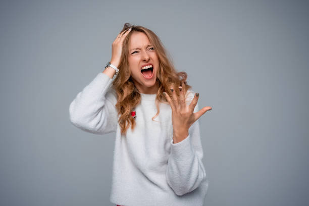 jeune femme irritée hurlant désespérément avec sa main sur la tête et la bouche grande ouverte, l’air en colère, porte un pull blanc, debout sur fond gris - mouth open women displeased anger photos et images de collection