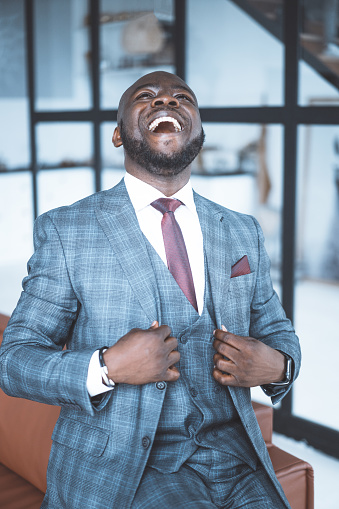 Successful Businessman in Formal Suit is Laughing with his Head Thrown Back. African Businessman Happy with a Successful Deal.Three-piece Suit. Close-up Portrait. Big Window Background . High quality photo