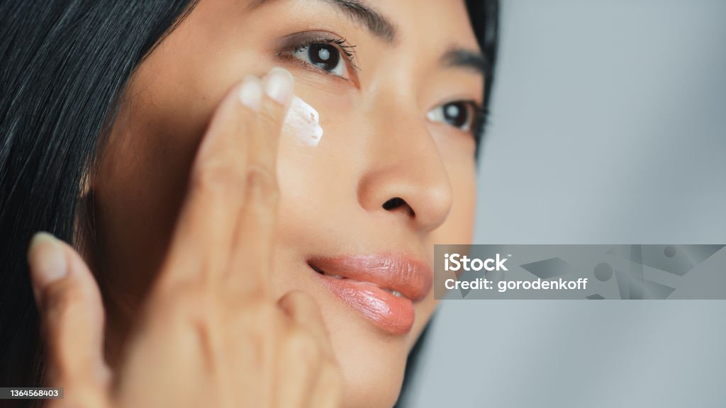 Beautiful Asian Woman Gently Touching Her Face, Looking Away. Young Adult Female with Lush Black Hair Makes Her Skin Soft, Clean with Natural Cosmetics Skincare Product. Close-up Portrait Face Cream Stock Photo
