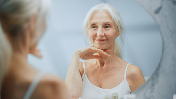 hermosa mujer mayor rutina de baño matutino, se mira en el espejo toca su cuello con el movimiento sensual de las manos. belleza natural femenina anciana. dignidad y gracia en la vejez. concepto para el cuidado de la piel - skin care blue eyes expressing positivity cheerful fotografías e imágenes de stock