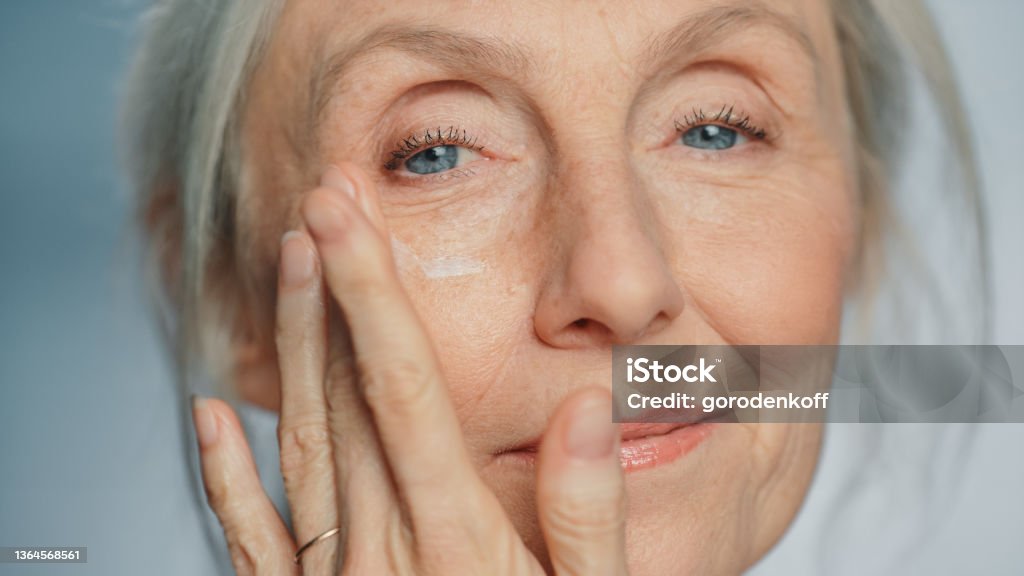 Portrait of Beautiful Senior Woman Gently Applying Under Eye Face Cream. Elderly Lady Makes Her Skin Soft, Smooth, Wrinkle Free with Natural anti-aging Cosmetics. Product for Beauty Skincare, Makeup Skin Stock Photo