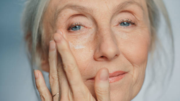 portrait of beautiful senior woman gently applying under eye face cream. elderly lady makes her skin soft, smooth, wrinkle free with natural anti-aging cosmetics. product for beauty skincare, makeup - skin care blue eyes expressing positivity cheerful imagens e fotografias de stock