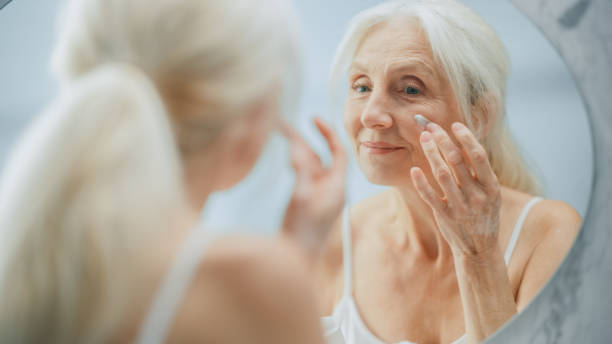 porträt der schönen älteren frau morgenroutine, blick in den spiegel sanft gesichtscreme auftragen. ältere dame macht ihre haut weich, glatt, faltenfrei mit natürlichen anti-aging-kosmetika, produkte - human face mature women make up women stock-fotos und bilder