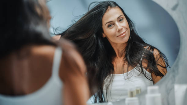 bella donna di mezza età guarda nello specchio del bagno tocca i suoi lussureggianti capelli neri, ammira il suo aspetto. concetto per la felicità, il benessere, la bellezza naturale, i prodotti biologici per la cura della pelle - healthy hair foto e immagini stock