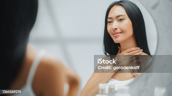 istock Beautiful Asian Woman Touches Her Perfect Soft Shoulder, Neck, Sensually Smiles in the Mirror. Happy Female Enjoying Her Beauty. Wellness Natural Cosmetic Skincare Products. 1364569275
