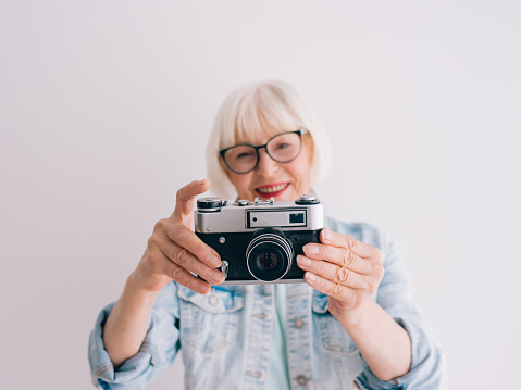 senior stylish woman with gray hair and in glasses and denim jacket taking pictures of flowers with film camera.