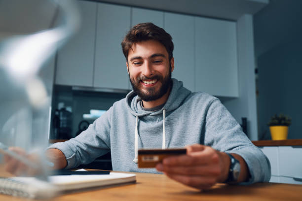 Smiling young man making online payments at home with credit card and laptop stock photo